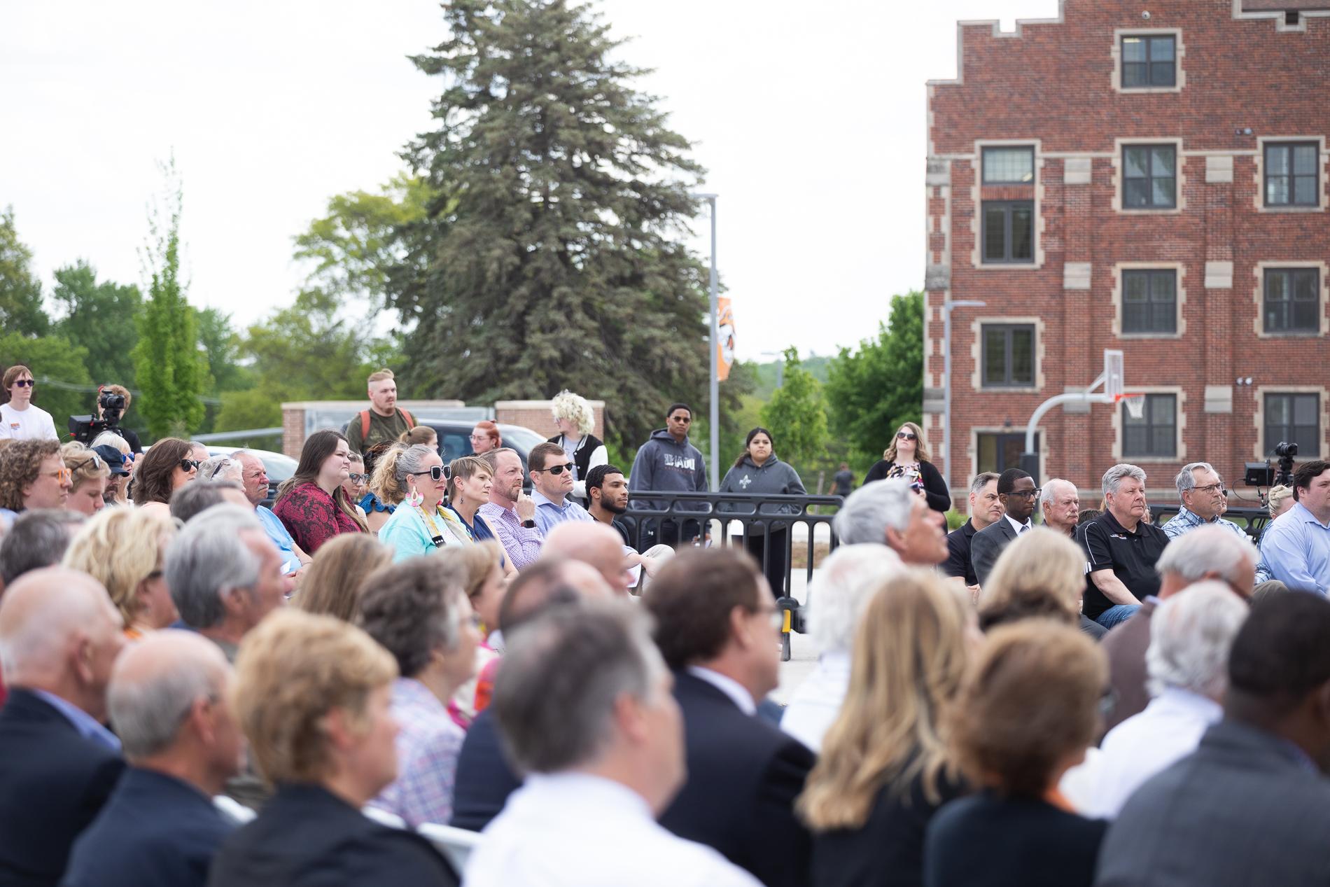 An audience is seated in rows, with several people standing behind them. In the background is a large brick building and trees. 