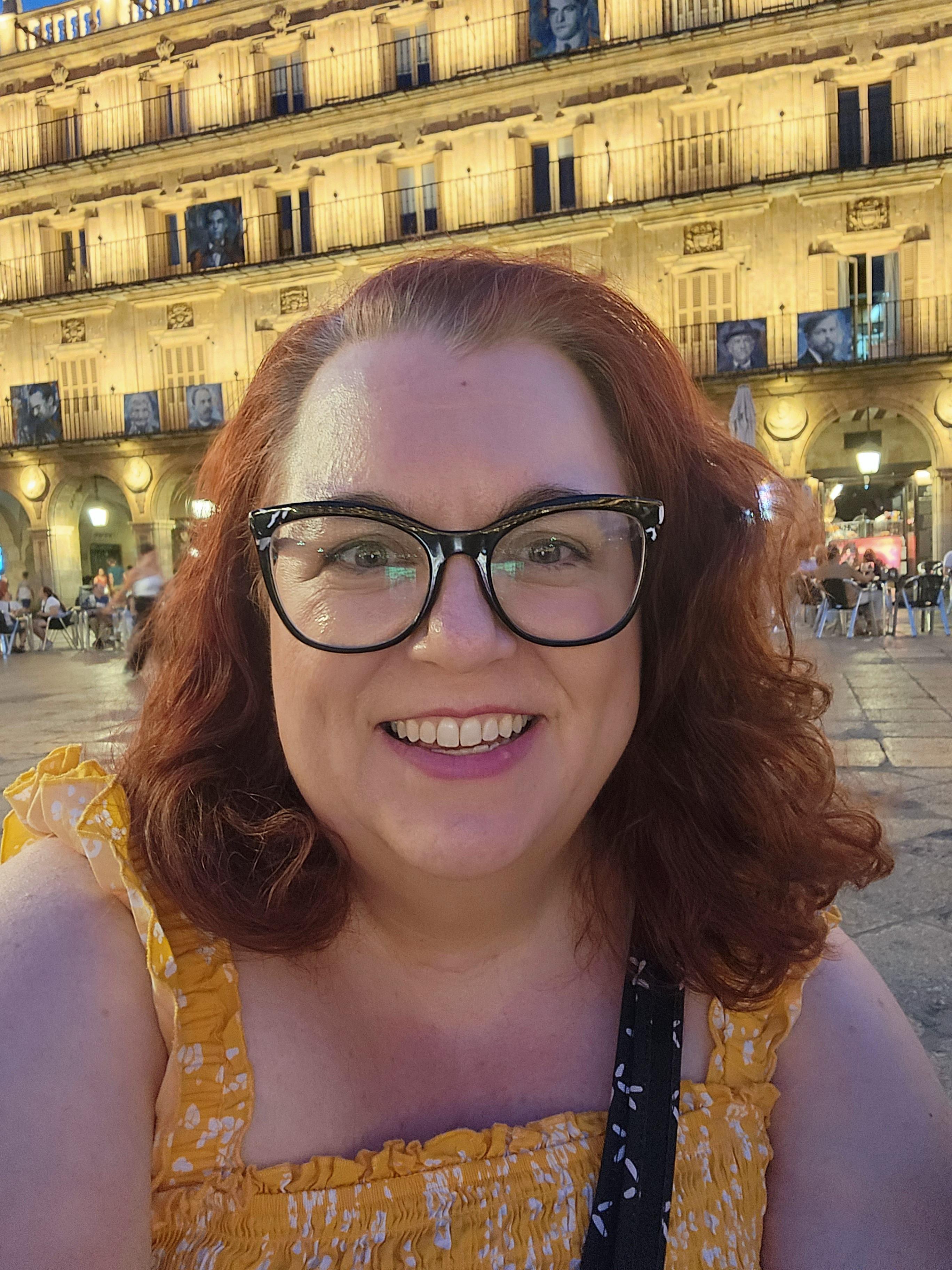 Melanie D'Amico takes a self-portrait while traveling abroad in Spain. She has curling, red hair and large, cat-eye glasses, and wears a yellow top.