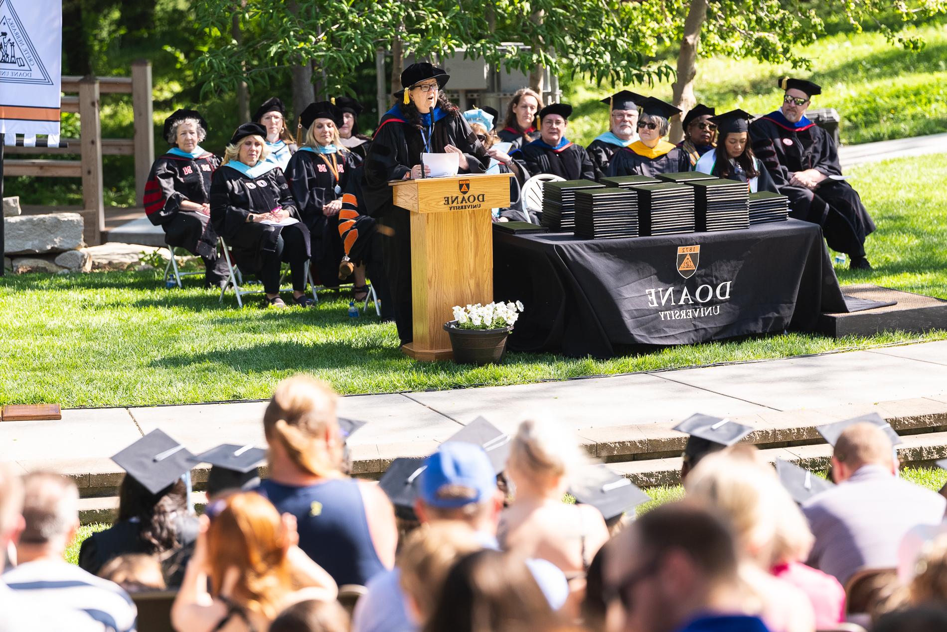 A woman in a black robe 和 cap speaks behind a podium, next to a table covered with thin, black folios. Behind her are more faculty members in black robes, 和 an audience is seated in front.