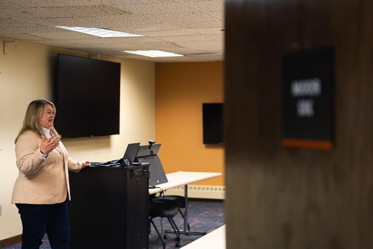 Dr. Jennifer Bossard在商学院技术教室的前面讲话, located in the Fred Brown Center in Lincoln. A partially closed door covers part of the image, while behind Bossard are two large televisions, a tablet and a cabinet with additional technology.